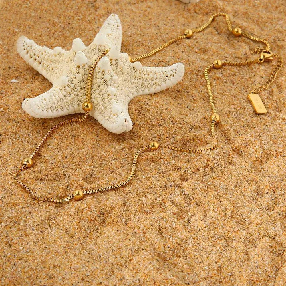 Schlichte Basic-Kette mit minimalistischem Design, liegend auf feinem Sand und Seesteinen am Strand. Die zeitlose Kette fügt sich harmonisch in die natürliche Umgebung ein und symbolisiert schlichte Eleganz.